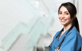Female caregiver in blue scrubs stands with arms crossed and stethoscope around her neck with a big simile on face in front of staircase in a home care agency.