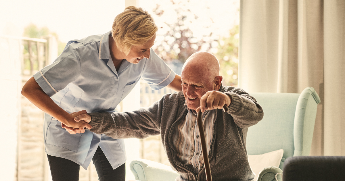 In-home caregiver assists elderly man with cane out of his chair while providing home care services