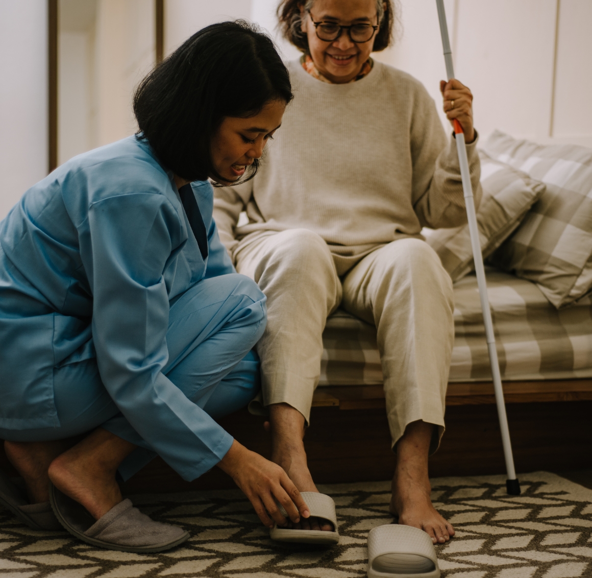 24 Hour Senior Care in Bradenton (1) A young Asian female caregiver sits on the floor helping an Asian senior woman put on her slippers, while the senior, seated on a sofa, looks down at her with a warm expression.