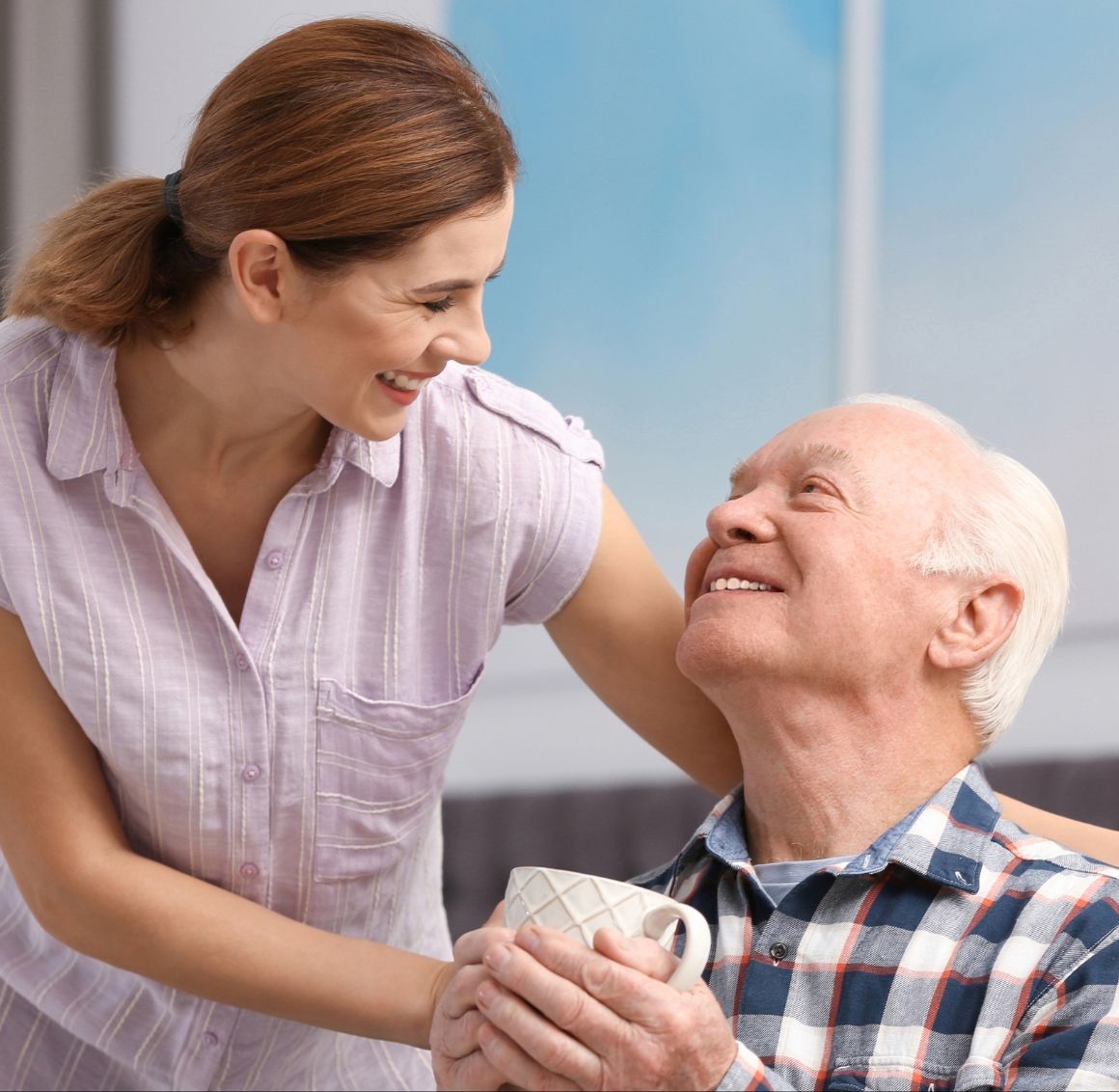 24 Hour Senior Care in Bradenton A female caregiver with her hair tied back places her hands on the shoulders of a senior man with grey hair while handing him a cup of tea, as he looks at her with a thankful expression.