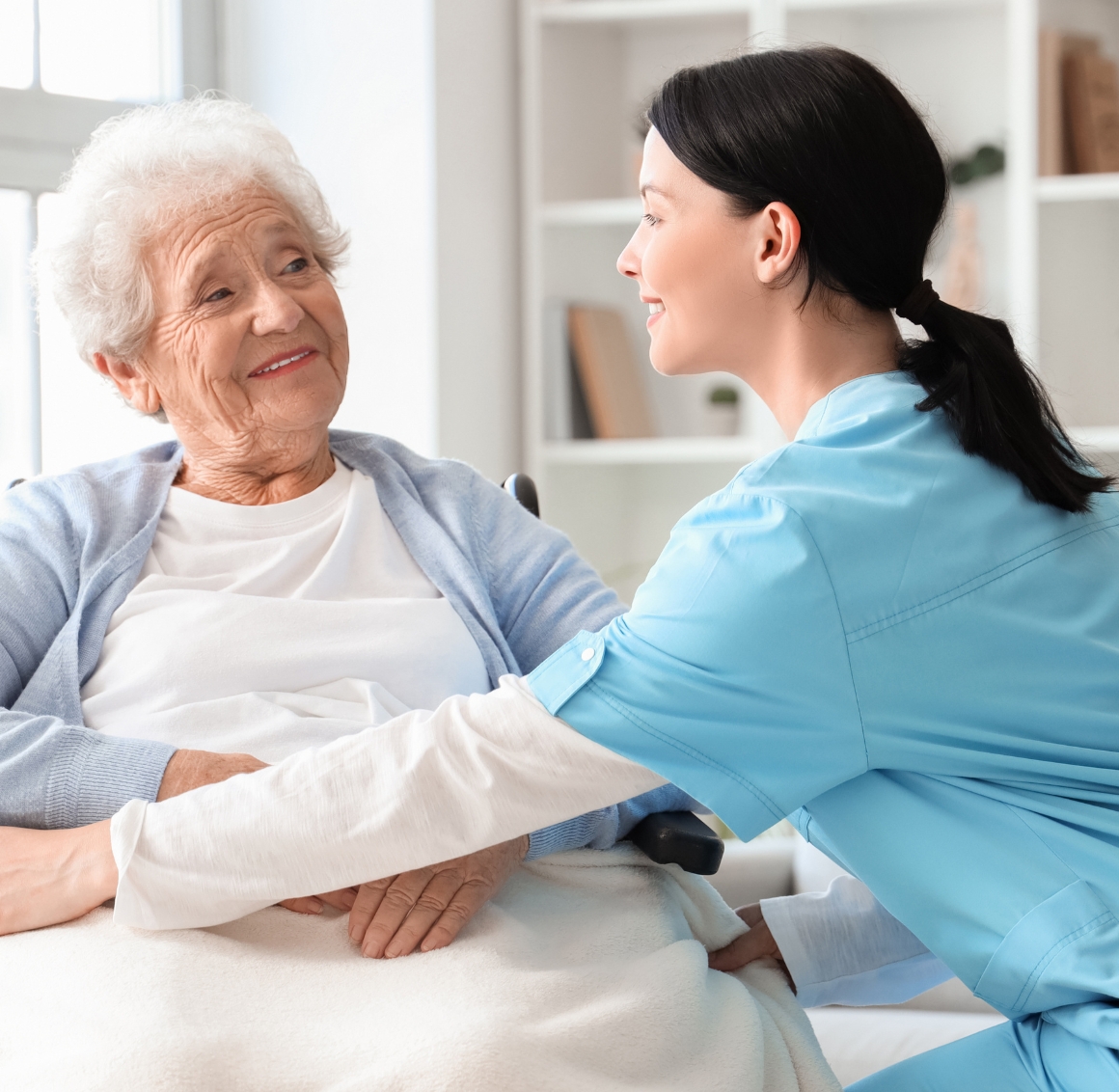 24 Hour Senior Home Care (1) A caregiver in blue scrubs smiles while assisting a senior woman in a wheelchair, representing compassionate 24 Hour Senior Home Care in Lakewood Ranch.