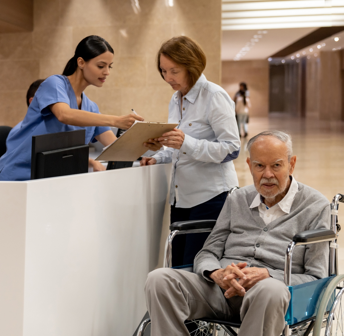A caregiver assists a senior man in a wheelchair at a hospital reception desk, representing supportive Hospital Companions in Lakewood Ranch.