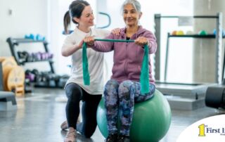 A care provider helps an older woman exercise with a resistance band and an exercise ball, representing how exercise can help with senior fall prevention.
