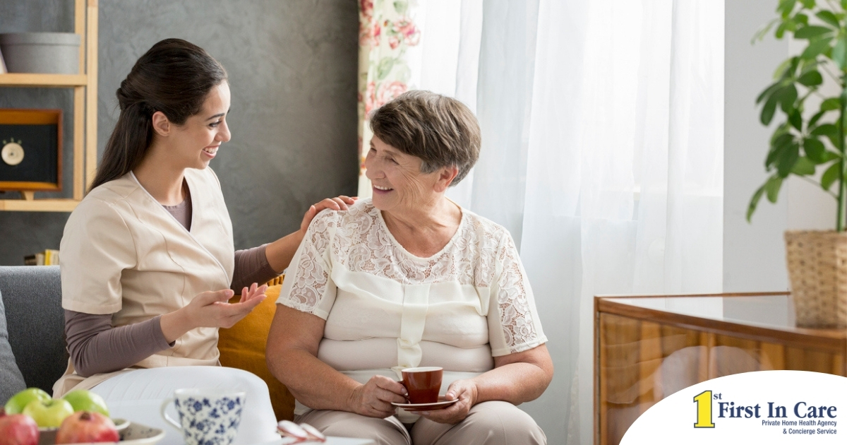 Smiling caregiver with her hand on an older woman’s shoulder as they share tea on the couch, showing home care vs assisted living